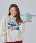 A woman in a cream sweatshirt holding a light green pot with purple handles and lid, smiling and winking against a light gray background