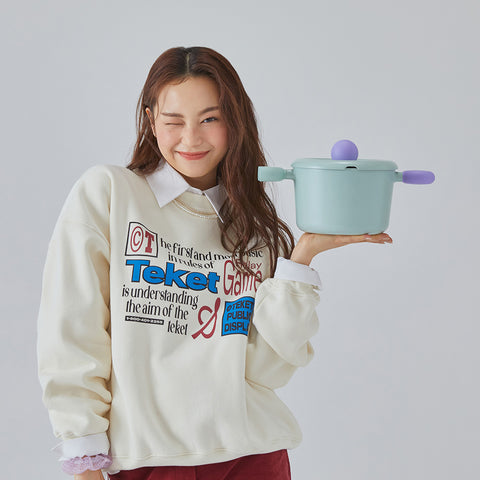 A woman in a cream sweatshirt holding a light green pot with purple handles and lid, smiling and winking against a light gray background