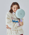 A woman in a cream sweatshirt holding a light green frying pan with a purple handle, smiling and posing against a light gray background