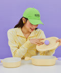 A woman in a yellow sweater and green cap, smiling as she holds the lid of a yellow oval pot with purple handles, sitting at a table against a purple background