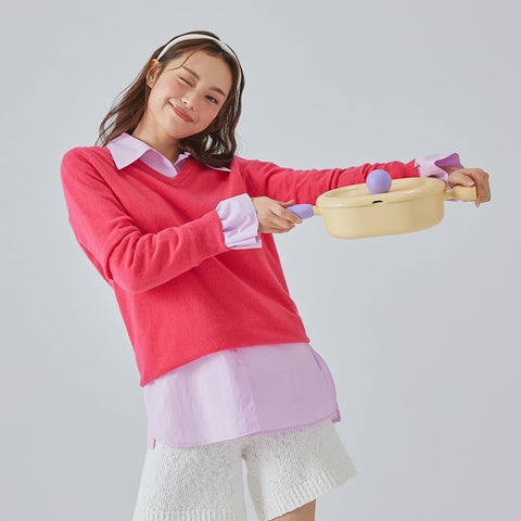 A woman in a cream sweatshirt holding a yellow frying pan with a purple handle, smiling and posing against a purple background