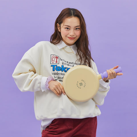 A woman in a red sweater and purple collared shirt, holding a yellow pot with purple handles, smiling and winking against a light gray background