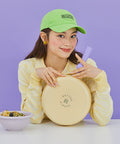 A woman in a yellow sweater and green cap, smiling as she holds a yellow frying pan with a purple handle, sitting at a table with a bowl of pasta, against a purple background