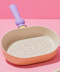 A pastel-colored grill pan with a light pink body and a purple handle, displayed on a pink background