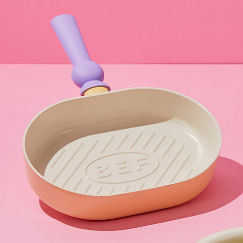 A pastel-colored grill pan with a light pink body and a purple handle, displayed on a pink backgroun