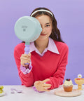 A woman in a red sweater and purple collared shirt, holding a light green frying pan with a purple handle in front of her face, sitting at a table with grapes and dessert items, against a purple background