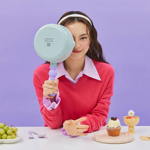 A woman in a red sweater and purple collared shirt, holding a light green frying pan with a purple handle in front of her face, sitting at a table with grapes and dessert items, against a purple background