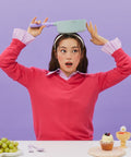 A woman in a red sweater and pink collared shirt, holding a light green pot with a purple handle on her head, sitting at a table with grapes and dessert items, against a purple background