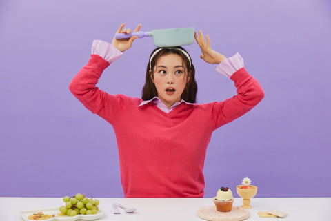 A woman in a red sweater and pink collared shirt, holding a light green pot with a purple handle on her head, sitting at a table with grapes and dessert items, against a purple background