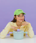 A woman in a yellow sweater and green cap, smiling as she poses with a light green pot with a lid and purple handles, sitting at a table against a purple background