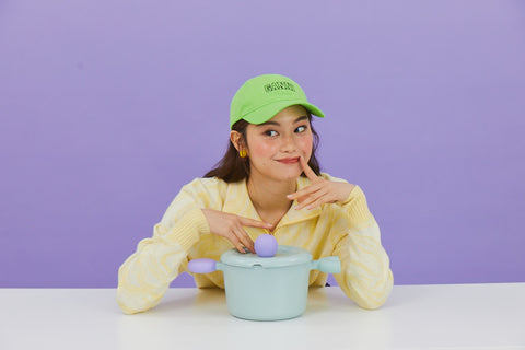 A woman in a yellow sweater and green cap, smiling as she poses with a light green pot with a lid and purple handles, sitting at a table against a purple background