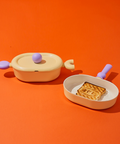 Two pastel-colored cookware items, including a yellow oval pot with a lid and purple handles, and a yellow grill pan with a purple handle, containing a grilled piece of bread with the 'BEF' logo, displayed on an orange background