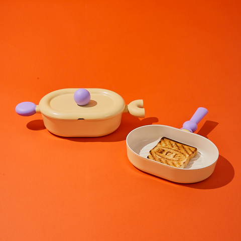 Two pastel-colored cookware items, including a yellow oval pot with a lid and purple handles, and a yellow grill pan with a purple handle, containing a grilled piece of bread with the 'BEF' logo, displayed on an orange background