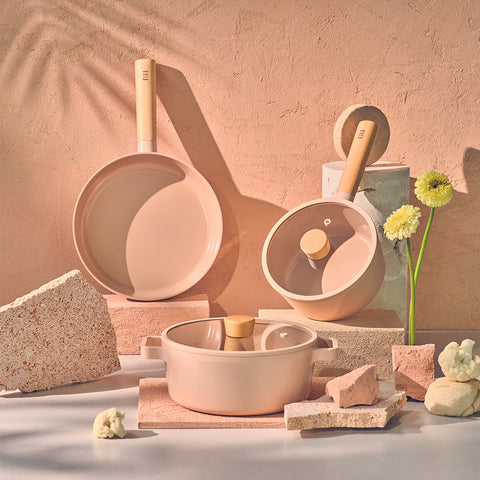 Aesthetic display of cooking utensils arranged on a pink-hued background with soft shadows. Includes a variety of pink and beige pots and pans, some with wooden handles, alongside decorative bricks and a small bouquet of yellow flowers for a touch of natural beauty.