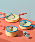 A neatly organized collection of Danish cookware on a vibrant orange surface. The set includes a yellow 8-inch casserole and a 7-inch saucepan with blue glass lids and red handles. An open 9-inch multi-pan displays a non-stick surface, hinting at its cooking efficiency. The cookware is accompanied by playful geometric kitchen accessories, emphasizing a modern and youthful kitchen aesthetic