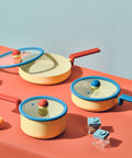 A neatly organized collection of Danish cookware on a vibrant orange surface. The set includes a yellow 8-inch casserole and a 7-inch saucepan with blue glass lids and red handles. An open 9-inch multi-pan displays a non-stick surface, hinting at its cooking efficiency. The cookware is accompanied by playful geometric kitchen accessories, emphasizing a modern and youthful kitchen aesthetic