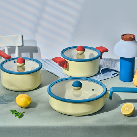 A serene kitchen scene showcasing Danish cookware on a pastel green surface. The image includes a 7-inch saucepan and an 8-inch casserole with a 9-inch multi-pan, all with glass lids and contrasting red and blue handles. Fresh lemons and a white towel are placed beside the cookware, accentuating the clean and fresh design of the high-quality cooking utensils