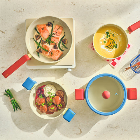An overhead view of a modern kitchen scene featuring Danish cookware. In the center is a 7-inch saucepan with a glass lid containing cooked salmon and garnished with herbs. Around it, there's a yellow multi-pan with red handles and a 9-inch casserole with a cream soup. The colorful cookware has distinctive red and blue handles, adding a pop of color to the neutral countertops
