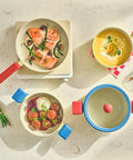 An overhead view of a modern kitchen scene featuring Danish cookware. In the center is a 7-inch saucepan with a glass lid containing cooked salmon and garnished with herbs. Around it, there's a yellow multi-pan with red handles and a 9-inch casserole with a cream soup. The colorful cookware has distinctive red and blue handles, adding a pop of color to the neutral countertops