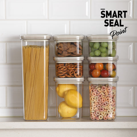 A neatly organized collection of transparent storage containers on a white shelf. The containers are filled with a variety of kitchen staples, including spaghetti, cookies, peas, cherry tomatoes, lemons, and cereal loops, each container labeled with 'Smart Seal Point' at the top