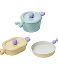 A set of three pastel-colored cookware items, including a light green pot with a lid, a yellow oval pot with a lid, and a yellow frying pan, all with purple handles, displayed on a white background