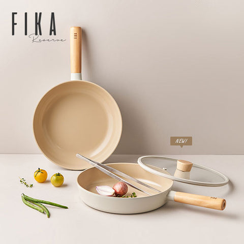Elegant kitchenware display from FIKA featuring a beige non-stick frying pan with a wooden handle and two matching pots with lids, one of which contains a wooden spoon. The cookware is arranged on a light cream surface, accompanied by fresh ingredients including green chili peppers, yellow cherry tomatoes, and garlic cloves, evoking a sense of fresh cooking. In the background, the 'FIKA Kitchenware' logo in a sophisticated font adds a branding element to the image