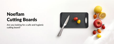 An advertisement for Noeflam Cutting Boards, featuring a dark grey cutting board on a white countertop. The board is adorned with a variety of colorful fresh produce including cherry tomatoes, a lemon, and a red apple, alongside a chef's knife. The text to the left of the board poses the question 'Are you looking for a safe and hygienic cutting board?' suggesting the product's key attributes