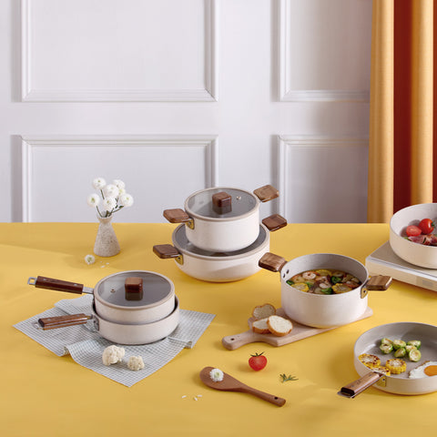 Alt text: "A colorful kitchen scene on a yellow tablecloth showcasing various white pots and pans with wooden handles. The cookware is arranged with a small vase of white flowers, fresh ingredients like garlic and tomatoes, and partially eaten dishes including soup and a salad plate. The background features elegant white paneling and a hint of golden drapery.