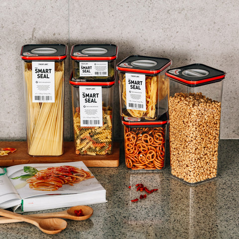 Assortment of four Smart Seal clear glass storage jars on a kitchen countertop, each filled with different dry foods. From left to right, the jars contain spaghetti, fusilli pasta, pretzel snacks, and oat flakes, with their airtight red lids and the Smart Seal label visible. Below the jars, there is an open cookbook with a picture of spaghetti with tomato sauce, a wooden spoon, and scattered ingredients including pasta and red chili flakes.